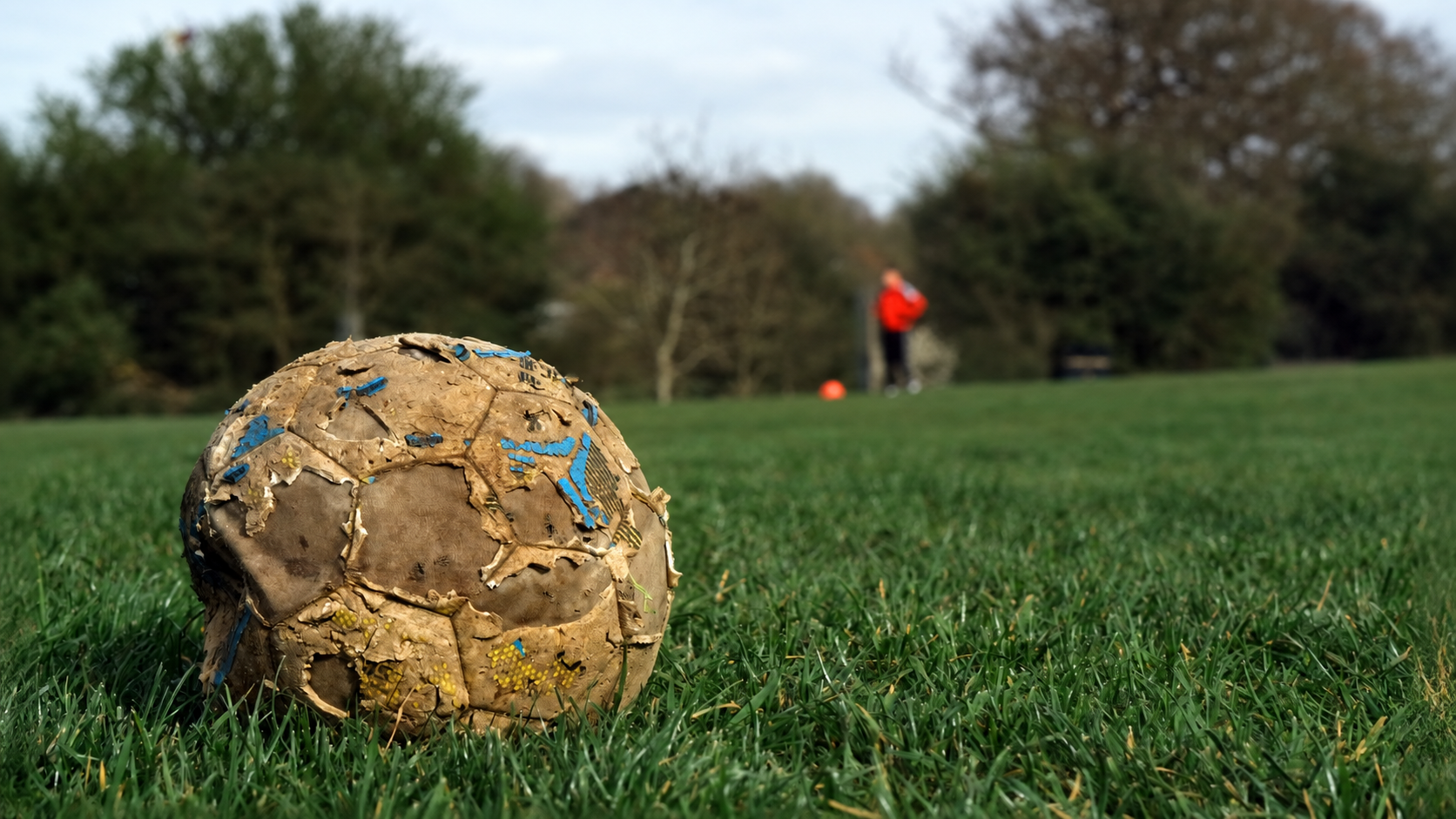 A heavily worn soccer ball resting on a grass field with a blurred figure in the distance
